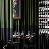 A styled interior scene featuring dark green and black velvet curtains, a wall-mounted wine rack filled with wine bottles, dining tables with glassware, dark wood chairs, and a large potted palm plant.