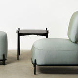 A modern lounge chair with a matching cylindrical ottoman and a small side table, all upholstered in Genre mist green fabric with black metal frames, set against a minimalist off-white wall and concrete floor.