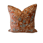 A square decorative throw pillow with a rust-orange background and an intricate floral pattern in cream, beige, and slate blue, shown against a plain white background.