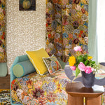A cozy, vintage-style bedroom scene featuring a daybed covered in a dense floral and botanical print fabric, styled with yellow and patterned accent pillows. The room is decorated with matching patterned wallpaper, curtains, and a rug, with a wooden side table holding a vase of pink and yellow flowers in the foreground.