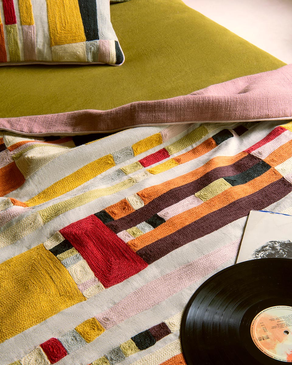 A close-up of a colorful patchwork textile, likely a throw blanket or quilt, draped over a green sofa with a pink pillow. A black vinyl record and a small card are partially visible in the foreground.