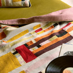 A close-up of a colorful patchwork textile, likely a throw blanket or quilt, draped over a green sofa with a pink pillow. A black vinyl record and a small card are partially visible in the foreground.