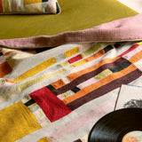 A close-up of a colorful patchwork textile, likely a throw blanket or quilt, draped over a green sofa with a pink pillow. A black vinyl record and a small card are partially visible in the foreground.