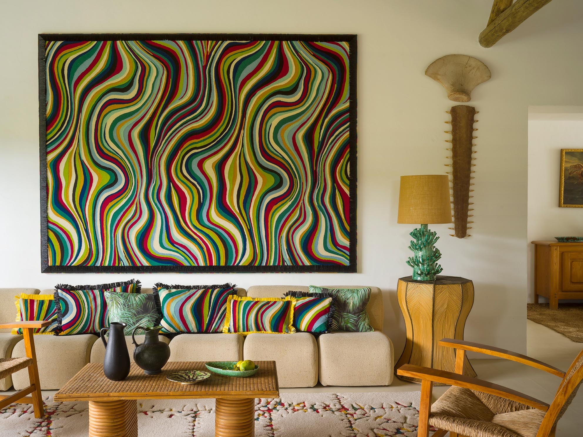 A large framed abstract wall art piece with bold wavy stripes in many colors hangs above a beige sofa in a styled living room. The room also includes patterned throw pillows, a woven coffee table, chairs, a side table with a lamp, and a tall decorative object on the wall.