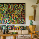 A large framed abstract wall art piece with bold wavy stripes in many colors hangs above a beige sofa in a styled living room. The room also includes patterned throw pillows, a woven coffee table, chairs, a side table with a lamp, and a tall decorative object on the wall.
