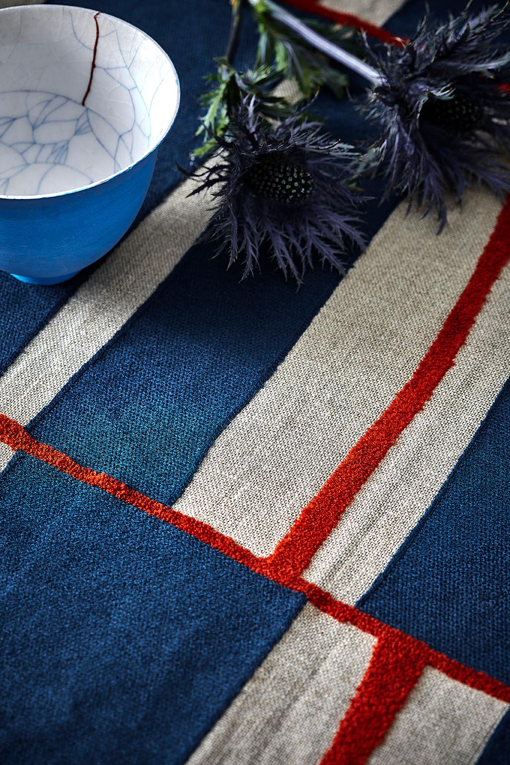 A close-up of a woven textile with broad navy and beige stripes and thin red lines, styled with a blue ceramic bowl and dark thistle flowers.