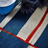 A close-up of a woven textile with broad navy and beige stripes and thin red lines, styled with a blue ceramic bowl and dark thistle flowers.