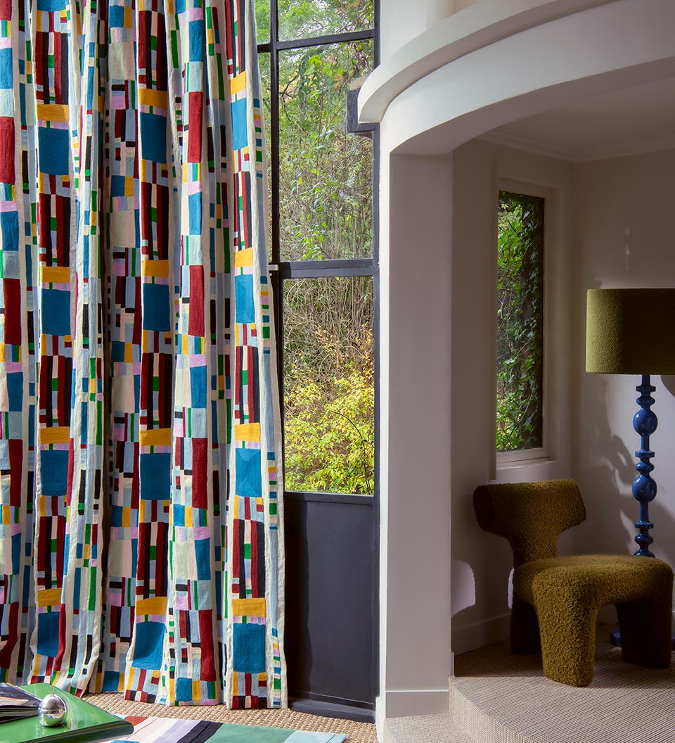 A tall window dressed with floor-length curtains featuring a bold, colorful geometric block pattern on a white base. The room setting includes a curved white wall, a textured olive-green armchair, a blue pedestal floor lamp, and a view of green trees outside.