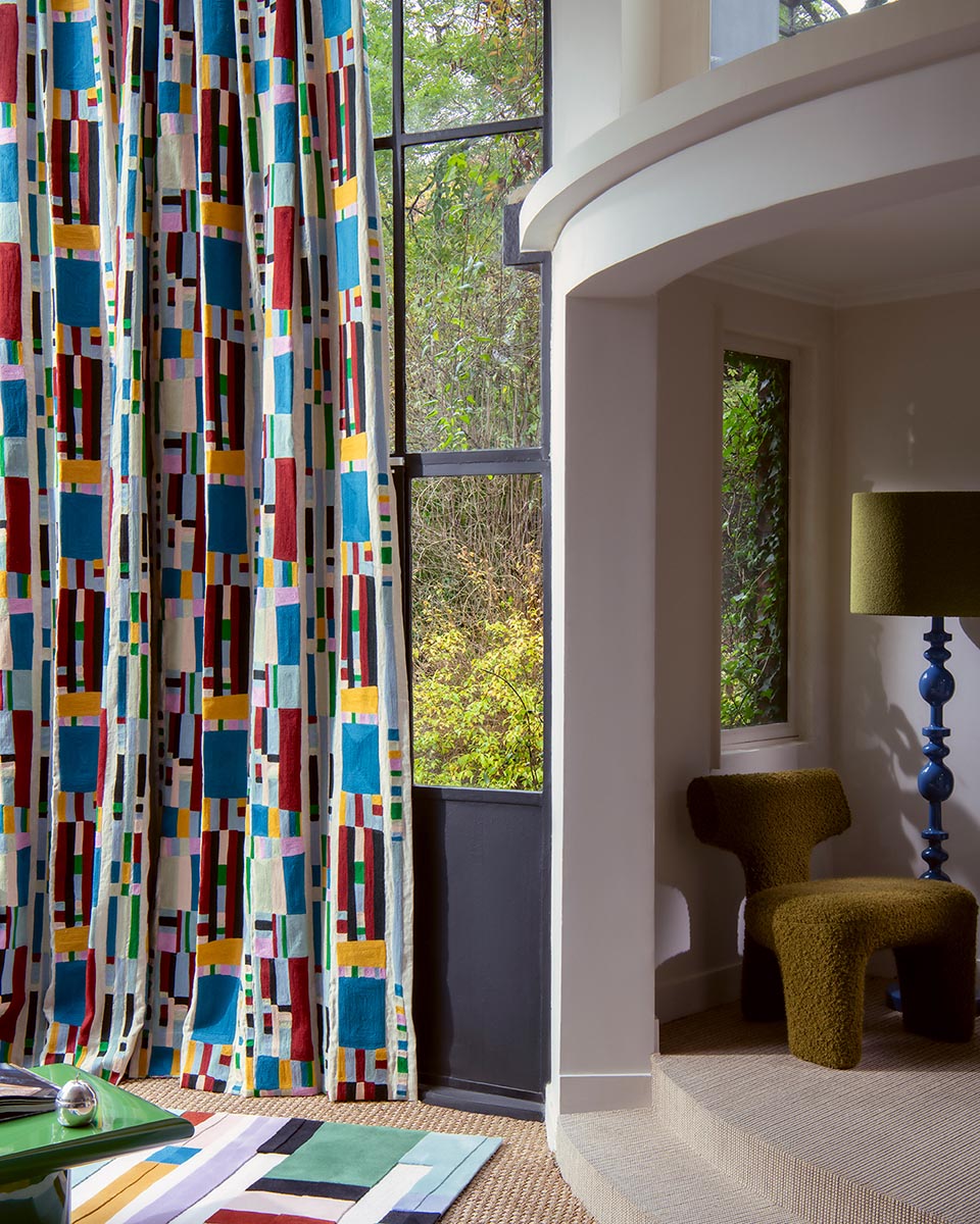 A tall window dressed with floor-length curtains featuring a bold, colorful geometric block pattern on a white base. The room setting includes a curved white wall, a textured olive-green armchair, a blue pedestal floor lamp, and a view of green trees outside.