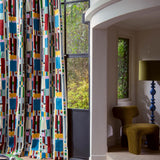 A tall window dressed with floor-length curtains featuring a bold, colorful geometric block pattern on a white base. The room setting includes a curved white wall, a textured olive-green armchair, a blue pedestal floor lamp, and a view of green trees outside.