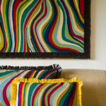 A colorful striped throw pillow with yellow fringe sits on a beige sofa, with a matching striped pillow and a large matching wall hanging above it.