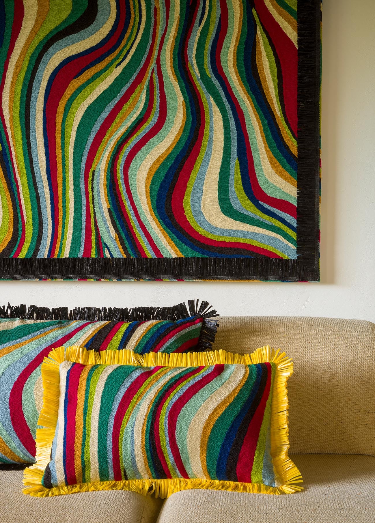 A colorful striped throw pillow with yellow fringe sits on a beige sofa, with a matching striped pillow and a large matching wall hanging above it.