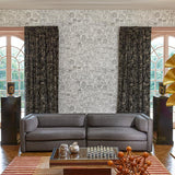 A styled interior room featuring a gray fabric sofa centered against a patterned wallpaper wall, with tall dark patterned curtains on either side of arched windows. A striped rug, low wooden coffee table, decorative objects, and a gold leaf floor lamp are also visible.