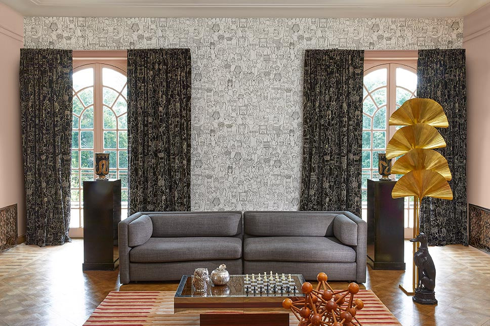 A styled interior room featuring a gray fabric sofa centered against a patterned wallpaper wall, with tall dark patterned curtains on either side of arched windows. A striped rug, low wooden coffee table, decorative objects, and a gold leaf floor lamp are also visible.