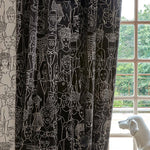 Black curtains with a white line-art illustration pattern of various figures and faces, hanging next to a window with a white dog statue in the foreground.