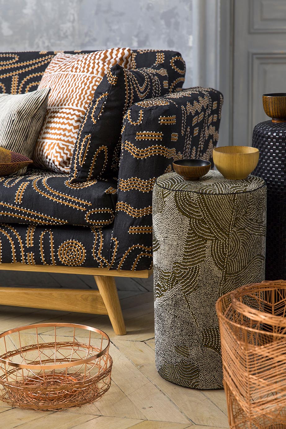 A styled interior scene featuring a dark upholstered armchair with a beige and brown abstract pattern, matching accent pillows, a cylindrical side table with a beige and gray pattern, and copper wire baskets on a stone floor.