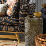 A styled interior scene featuring a dark upholstered armchair with a beige and brown abstract pattern, matching accent pillows, a cylindrical side table with a beige and gray pattern, and copper wire baskets on a stone floor.